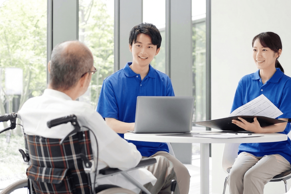 車椅子に座る高齢男性と、青い制服のスタッフ2人がテーブル越しに笑顔で会話しながら相談している介護の場面
